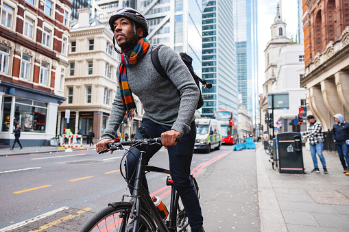 An adult black male cycling to his office in London. He is wearing a helmet for safety. The man looks focused and careful as he is biking. He is located on a beautiful city street with various architecture styles.
