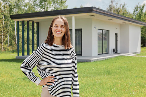 Dark haired smiling standing near building new house on green lawn keeps hand on hip smiling to camera wearing striped shirt having positive emotions.