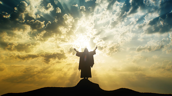 Silhouette of Jesus praying with raised hands on a mount with mystic clouds behind Him.