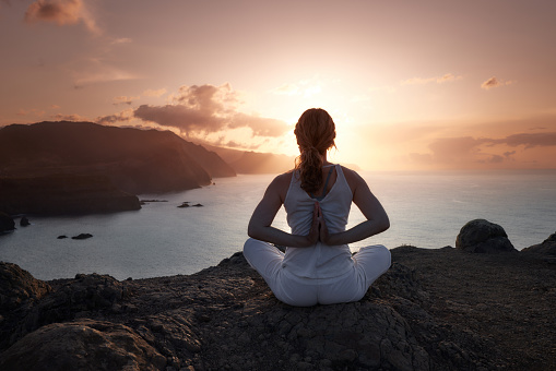 Back view of athletic woman doing Yoga relaxation exercises on a mountain above the sea at sunset. Copy space.