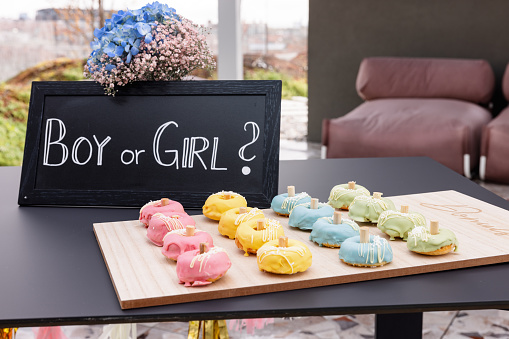 A table with delicious sweets and a sign 'Boy or Girl?' before a gender reveal party. The guests haven't arrived yet and there is no people. The doughnuts are cute and colorful. A beautiful idea for gender reveal decorations for an outdoor celebration in the summertime.