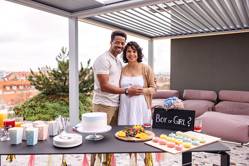 A happy diverse adult couple smiling at the camera and posing for a cute photo. They are both holding the pregnant belly and look happy. The couple is standing by a table that is decorated with cute gender-reveal decorations and is full of delicious fresh fruits and desserts.