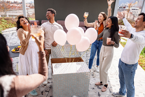 Excited and happy expectant couple finding out the gender of their baby by opening a box full of balloons. The balloons are pink indicating the couple is about to have a baby girl. They are surrounded by supportive diverse close friends that are cheering and clapping. They are located on a rooftop of a building.