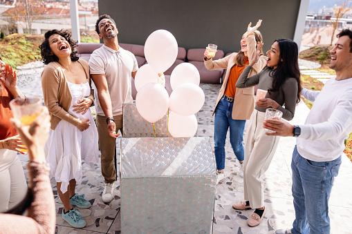 A group of diverse close friends smiling and cheering as they find out the expectant couple is about to have a baby girl. The balloons inside the box revealed the gender of the baby and since they were pink they are having a girl. A beautiful heartfelt and memorable moment for the couple and their closest friends. They are gathered outdoors on a beautiful rooftop.