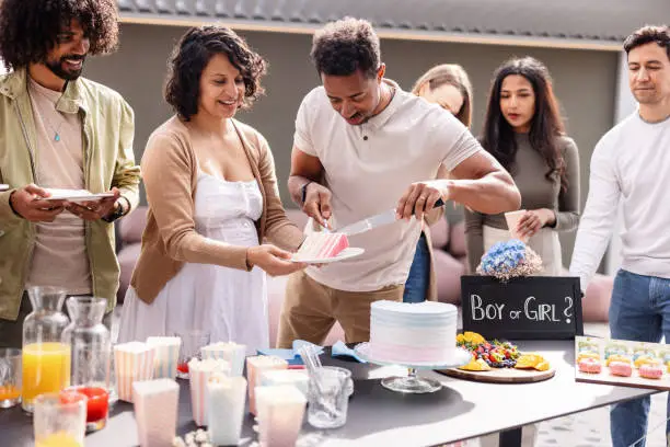 A multiracial expectant couple serving delicious cake to the diverse gender reveal party guests. They are celebrating having a girl on a rooftop terrace on a sunny spring day. Everyone looks happy and excited.