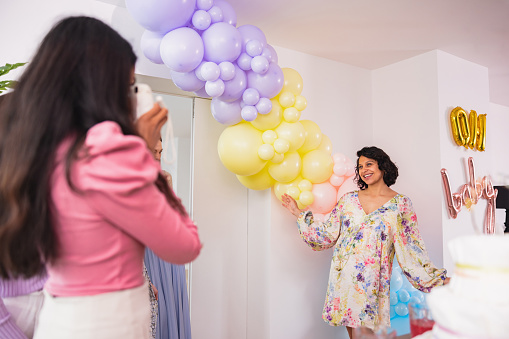 A happy Hispanic mom-to-be in a floral dress posing for a photo at her baby shower where they are celebrating her baby girl. She is surrounded by her supportive female friends. They are located in her modern apartment which has been decorated with colourful and vibrant decorations and balloons. Everyone is smiling and looks happy.