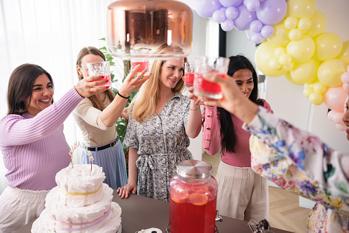 A multiracial group of female friends toasting and raising their glasses at a baby shower celebration. The female friend group is gathered around a table full of delicious party food and desserts. They are toasting to their pregnant friend and her baby girl. A beautiful heartfelt moment.