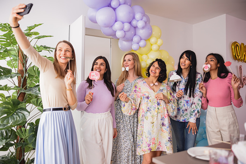 A cheerful group of multiracial female close friends taking funny photos with photo props at a baby shower celebration. They are smiling while taking cheerful photos together to commemorate the moment. Fun baby shower activities for friends.
