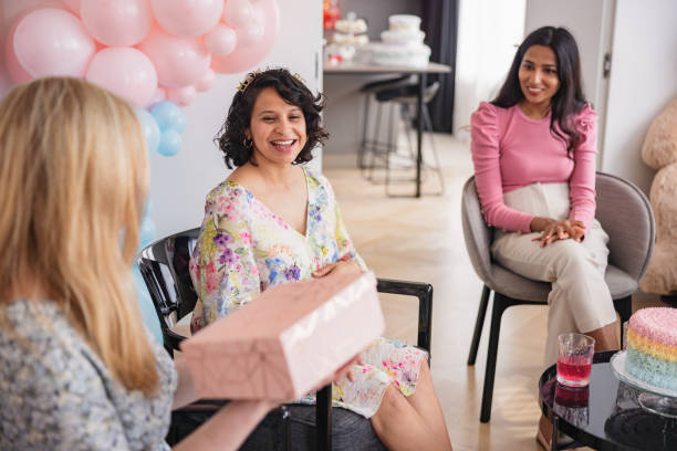 Gift Giving During A Surprise Baby Shower Celebration A Hispanic expectant mother receiving thoughtful gifts from her closest friends at a baby shower celebration. She looks expected and is smiling. They are all gathered together in a fancy apartment that is decorated with beautiful baby shower decorations and balloons. cute pregnant woman with gift stock pictures, royalty-free photos & images