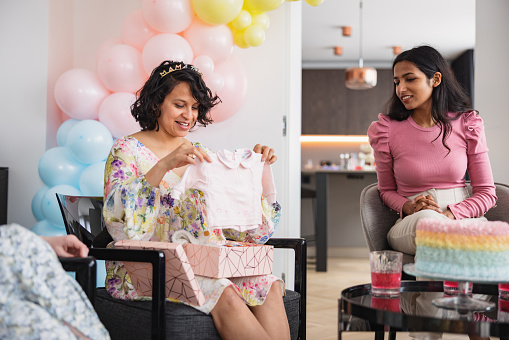A Hispanic mother-to-be smiling while opining the gifts her friends gave her at the baby shower. She looks happy and exited when seeing what they got her. Beautiful heartwarming moments at a celebration.