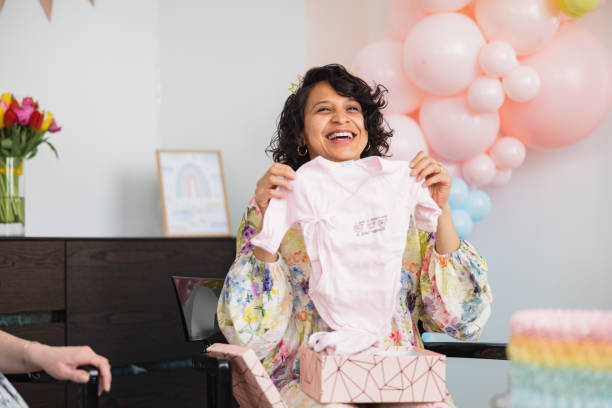 Happy Expectant Mother Opening Cute Gifts At A Baby Shower Celebration At Her Modern Apartment A Hispanic mother-to-be smiling while opining the gifts her friends gave her at the baby shower. She looks happy and exited when seeing what they got her. Beautiful heartwarming moments at a celebration. cute pregnant woman with gift stock pictures, royalty-free photos & images