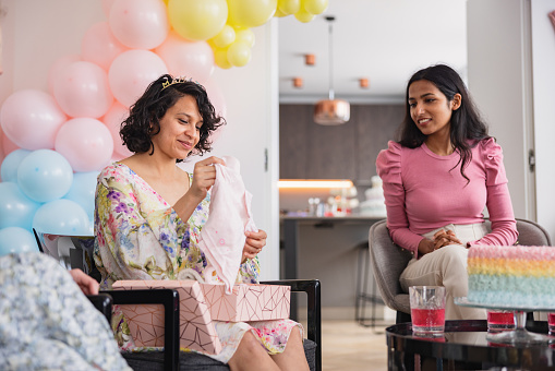 A happy young pregnant woman opening presents at a baby shower party planned by her best friends. They are gathered around the young mother-to-be while she is going over her gifts. There is a delicious cake and various desserts on the table. Exciting activities at a baby shower.