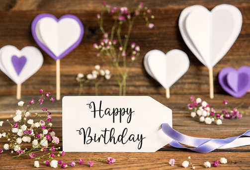 Label With English Text Happy Birthday. Purple And Lilac Decoration And Spring Flower Arrangement. Heart Symbols With Wooden Background.