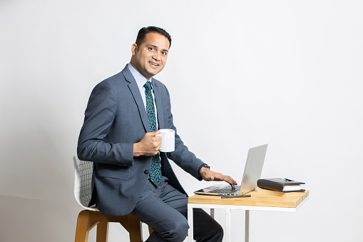 Portrait of young Indian business man wear suit using laptop while holding coffee mug in hand, Asian corporate guy working on computer sitting on chair looking at camera isolated on white studio background