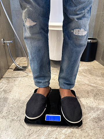 Stock photo showing close-up view of unrecognisable person, wearing denim jeans and slippers, standing on a pair of weighing scales in a luxury hotel bathroom with marbled effect patterned floor and wall tiles. Dieting and eating disorder concept.