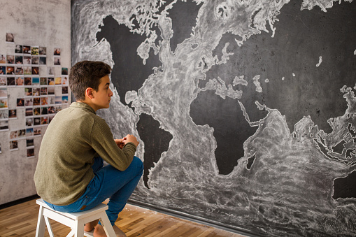 Teenage boy sitting in front of world map painting with chalk on a blackboard wall in the room