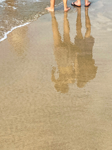 Stock photo showing a heterosexual couple strolling along the water's edge of a sandy beach, carrying shoes in their hands.