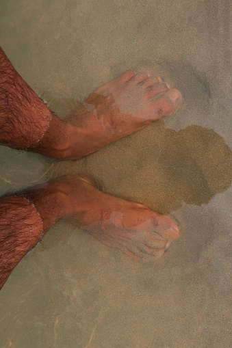 Stock photo of elevated view of barefoot man with feet standing ankle deep in sea water on beach.
