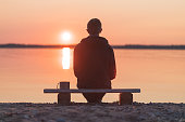 A man is sitting on a wooden bench on the shore of the sunset sea.