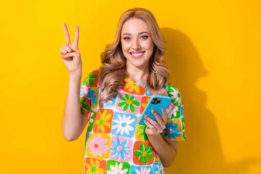 Photo of cute friendly woman dressed flower print t-shirt typing sms modern device showing v-sign isolated yellow color background.
