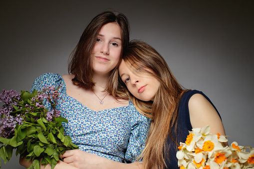 Portrait of two beautiful teenage girls standing close to each other, looking at camera and holding flower bunches, studio shot