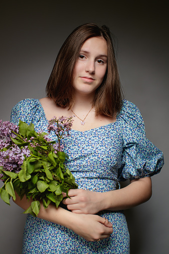 Portrait of beautiful teenage girl with brown hair in spring dress looking at camera and holding bunch of lilac, studio shot