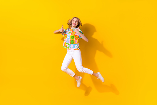 Full length photo of cool cheerful woman dressed flower print t-shirt jumping high showing two thumbs up isolated yellow color background.
