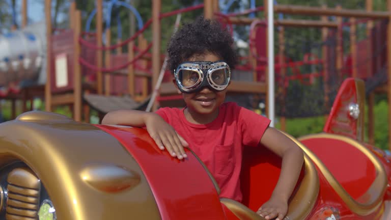 Portrait of adorable African-American-American toddler boy wearing pilot goggles sitting in toy plane on playground