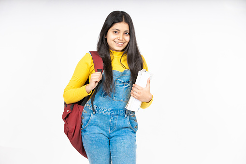 Portrait of happy indian teenager college or school girl with backpack holding books, isolated on white background. Smiling young asian female kid looking at camera.