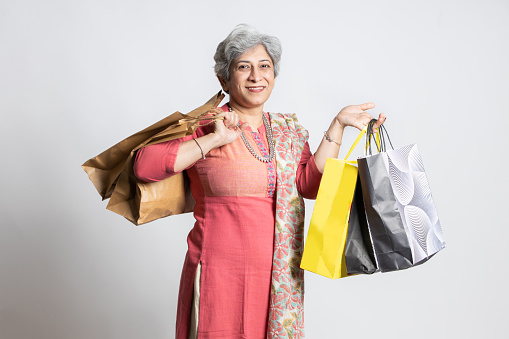 Happy indian senior mature woman holding shopping bags. elderly female shopper wearing traditional outfit, isolated white studio background, black Friday deal, diwali festive discount sale.