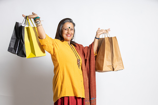 Happy excited indian senior mature woman holding shopping bags. elderly female shopper wearing traditional outfit, isolated white studio background, black Friday deal, diwali festive discount sale.
