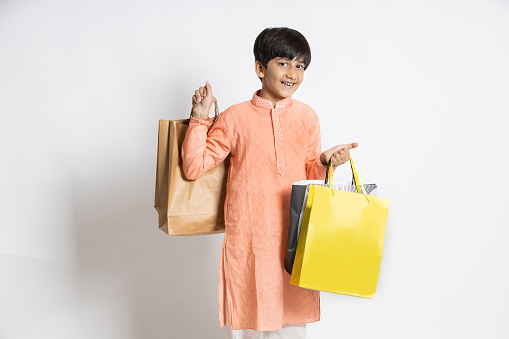 Happy little indian kid holding shopping or gift bags. male child shopper wearing ethnic kurta outfit, isolated over white studio background, black Friday deal, discount. festival sale.