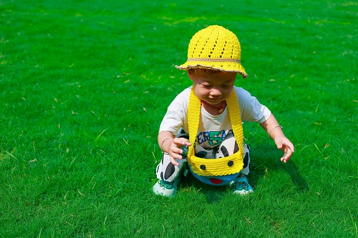 A cute toddler wearing a bright yellow bucket hat sits in the grass.