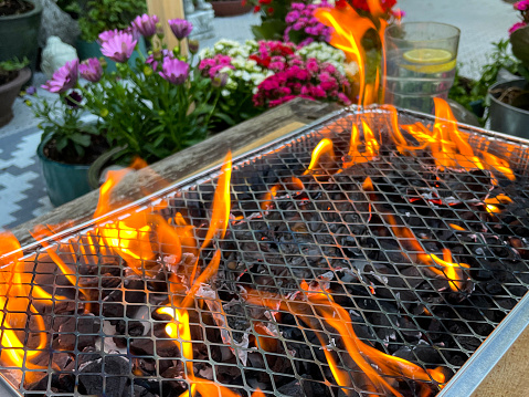 Stock photo showing close-up view of orange flames on charcoal briquette coals, just after the charcoal and small fire lighter paper have been lit with a long BBQ lighter tool. This disposable tinfoil tray BBQ is pictured burning away in a back garden, as it gets ready to cook lots of tasty sausages, burgers and kebabs.