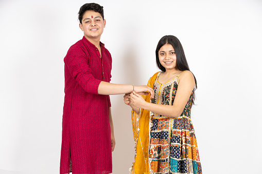 Portrait of happy indian teenage girl tying rakhi on young brother's hand occasion of rakshabandha or bhi dooj isolated on white studio background. both wearing traditional festive cloths.