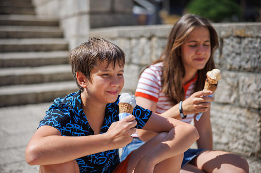 Teenage girl eating ice cream with her brother, sitting on stone stairs in the sun and eating ice cream, summer vacation in the town of Rab on Rab Island