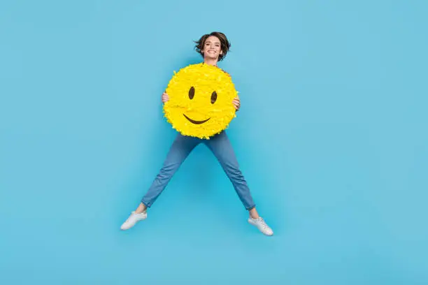 Photo of Full length photo of cute adorable lady wear denim jeans jumping high holding smiley pinata isolated blue color background