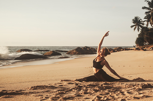 Young woman does yoga for healthy lifestyle on sea beach sitting on splits. Female performance sports exercises to restore strength and spirit. Yoga position on tropical climate