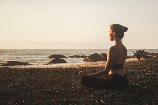 Young woman sitting in lotus position and does yoga for healthy lifestyle on ocean beach. Female performing sports exercises to restore strength and spirit. Yoga position on seashore