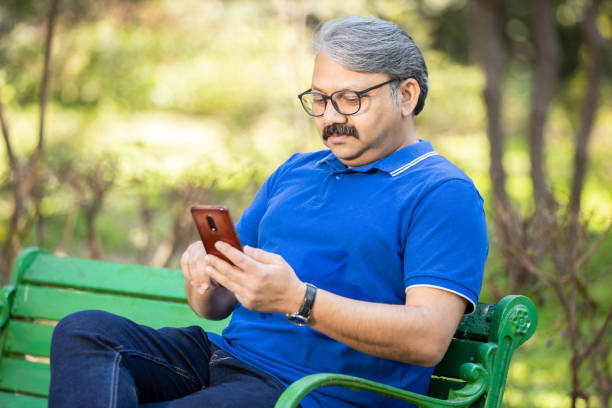 Happy Indian or asian senior man using chatting on smart phone while sitting on the bench at park , Old grey hair male wearing glasses relaxing using mobile phone outdoor, social media & technology.