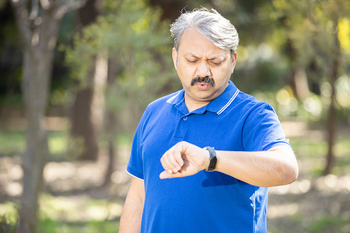 Senior Indian grey-haired man standing at park looking at the watch time worried, old male person afraid of getting late.
