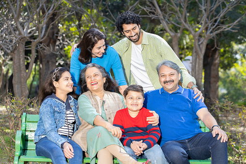 Happy indian family sitting together on a bench in a park laughing. Asian senior and young couple with their kids wearing casual cloths having fun, enjoying picnic holiday outdoor. selective focus