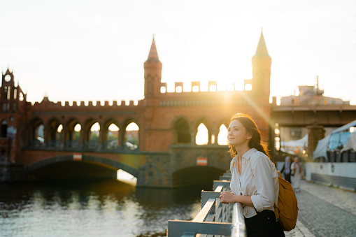 Woman standing at sunset and looking at scenic view of Berlin city and Oberbaumbrucke bridge. She is happy and carefree