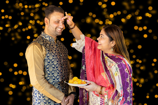 Young indian couple holding poja thali with full of sweets celebrating Diwali festival isolated on festive bokeh lights background.