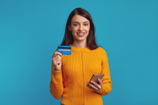 Portrait of a smiling casual girl in orange sweater holding mobile phone and plastic credit card isolated over blue background