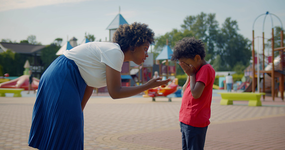 Side view of african strict mother talking to naughty son in park. Portrait of young woman scold preschool crying boy for bad behavior on playground