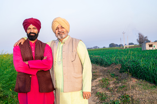 Portrait of happy punjabi srdar father and son wearing kurta and pagdi turban standing together at agriculture field.