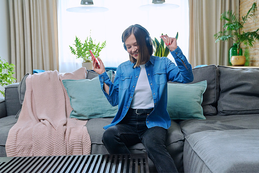 Young pretty woman in headphones with a smartphone enjoying music, having fun, with closed eyes, dancing, sitting on the sofa in the living room at home. Youth, fun, music, lifestyle concept