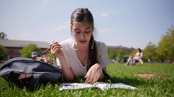 Portrait of female student in earphones study online outdoors. Young caucasian woman having online class writing notes lying on lawn in park