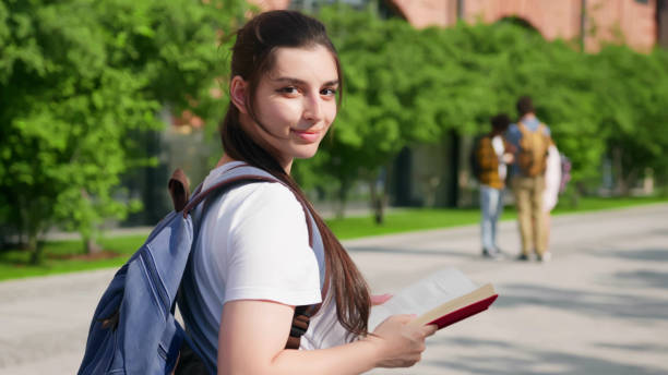 istock Happy university student walking with book and looking back at camera outdoors 1500863976